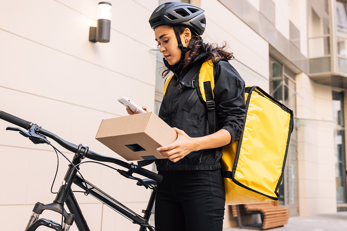 Female messenger wearing cycling helmet and backpack looking on mobile phone while holding a parcel