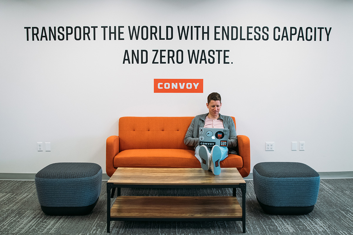 Convoy team member sitting on a couch working on a laptop with text above him on the wall that says "Transport the world with endless capacity and zero waste"
