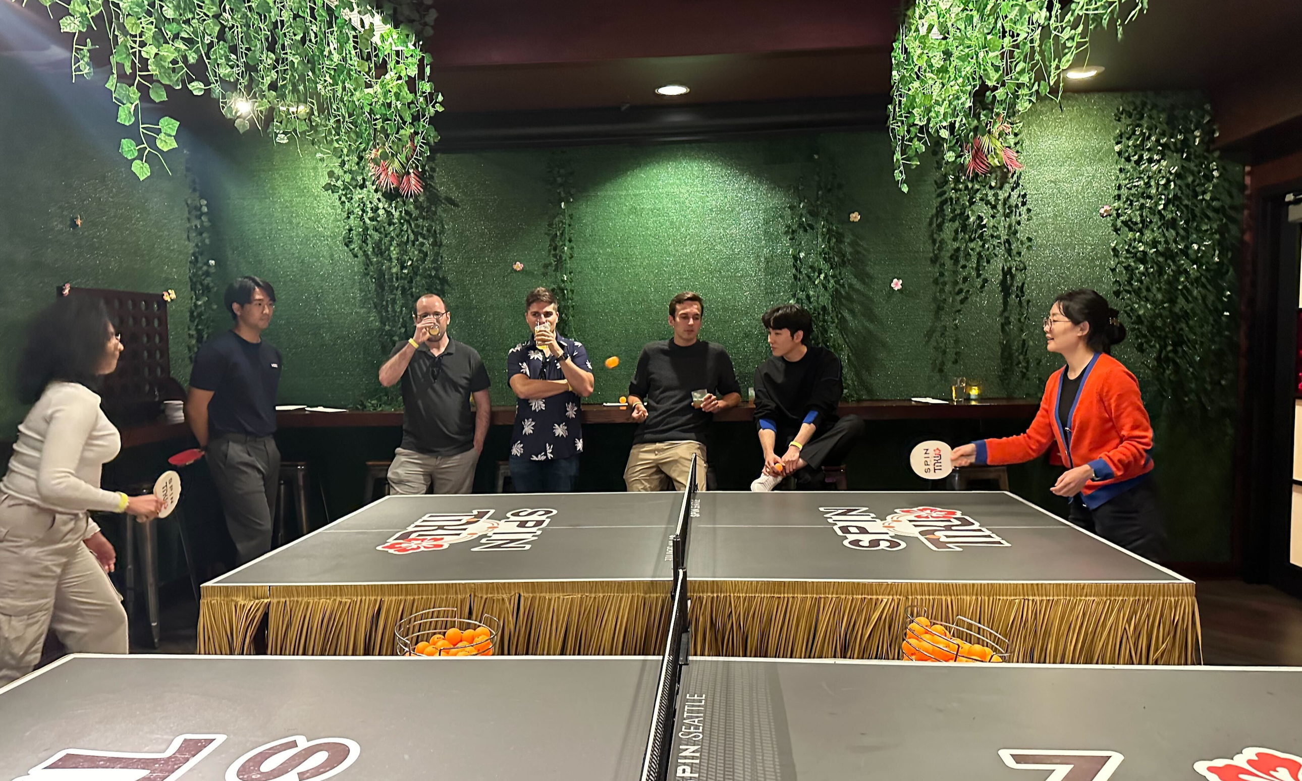 A group of Remitly employees play ping pong in a game room lined with hanging plants. 