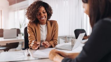 Woman sits at a desk in front of another woman, explaining something on a laptop that's between them