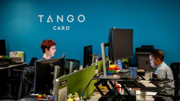 Two Tango Card employees at work under a blue wall and the Tango Card logo.