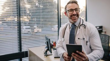 A male doctor smiles while looking off camera and holds a tablet