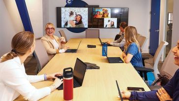 Meeting at a conference table with screens on wall showing Zoom participants with pets, team laughing