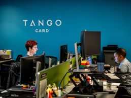 Two Tango Card employees at work under a blue wall and the Tango Card logo.